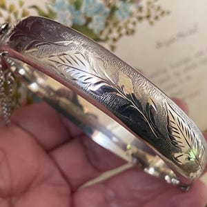 以下が含まれることがあります： 彫刻された葉のデザインが施された、銀色のヒンジ付きバングルブレスレット。 ブレスレットには安全チェーンとブラッシュ仕上げが施されています。 クラスプは閉じられています。 クラシックなジュエリーです。