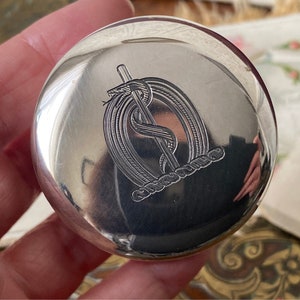 Könnte beinhalten: Ein runder Kompakt aus Silber mit einem gravierten Bild einer Schlange, die sich um einen Stab windet, ein Symbol, das mit Medizin in Verbindung gebracht wird.