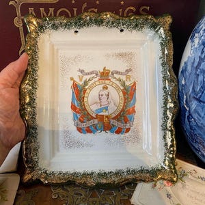 May include: A white square ceramic plate with a gold and green trim. The plate features a central emblem with a portrait, flags, and text. The plate is held by a hand.