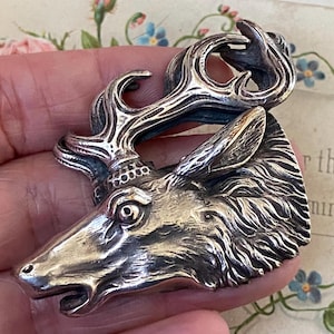 May include: A silver-toned metal brooch shaped like a deer head with detailed antlers. The piece features a textured finish that highlights the animal's fur, ears, and facial features. The deer is depicted in profile, facing left, with a prominent eye.