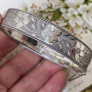 Może przedstawiać: Srebrzysta, otwierana bransoletka typu bangle z wygrawerowanym wzorem kwiatowym, polerowanymi płatkami i teksturowanymi liśćmi. Bransoletka jest wykończona skręconą krawędzią w stylu liny na górze i na dole oraz posiada metalowe zapięcie.