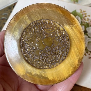 May include: A circular, light brown, polished horn or resin disc featuring a central, inset bronze-coloured metal medallion. The medallion displays an intricate embossed floral pattern surrounding a central shield and the letters H.P. Co.