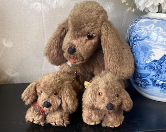 May include: Three brown plush dog toys. The largest dog has long floppy ears and a black nose. Two smaller dogs sit in front of the larger dog. One has a red ribbon around its neck. The dogs are on a black surface.