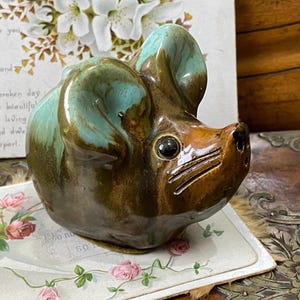 May include: A ceramic mouse figurine featuring a glossy brown body with a smooth, light turquoise glaze on the upper ears and head. The mouse has a pointed snout with three incised whisker lines on each side and a single dark, round eye.