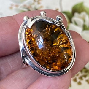 May include: A sterling silver ring featuring a large, oval-shaped, faceted amber gemstone with deep golden and orange hues. The metal setting includes smooth, curved silver bands and three small, polished silver spheres at the top edge.