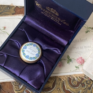May include: A small, round, gold-rimmed trinket box with a blue and white forget-me-not flower design. The box lid features the words "Forget me not" in black lettering. The box is in a blue velvet box.