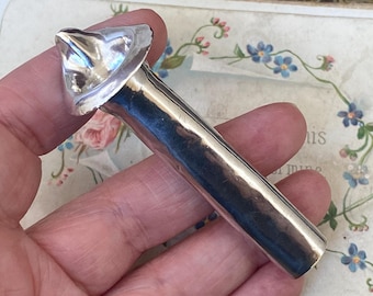 May include: A polished silver-toned metal tool featuring a cylindrical handle and a pointed, conical top section. The object has a smooth, reflective surface and a distinct shape that tapers to a point at one end, resembling a small citrus reamer.