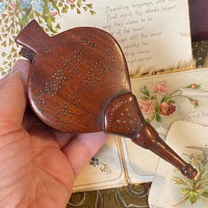 May include: A vintage wooden bellows, featuring a dark brown finish and brass embellishments. The rounded body is decorated with small brass studs. The bellows has a long, tapered nozzle. The object is held in a hand.