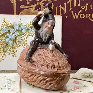 Puede incluir: Una caprichosa figura de gnomo de cerámica posada sobre una caja de baratijas con forma de nuez. El gnomo, vestido de marrón y negro, sostiene un pequeño martillo. La caja es de color marrón claro. Tarjetas decorativas y un libro en el fondo.