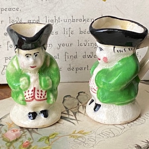 Puede incluir: Dos pequeñas figuritas de cerámica de personas que llevan chaquetas verdes y sombreros negros. Las figuritas están pintadas con detalles blancos, rojos y negros. Las figuritas tienen la forma de personas que llevan ropa tradicional.