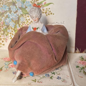 Puede incluir: Una muñeca de porcelana vintage sentada en un cojín de terciopelo malva. La muñeca tiene una peluca blanca con un lazo rojo, un vestido azul y una cara pintada. El cojín está adornado con dos cuentas azules. La muñeca está sobre una superficie con estampado floral.