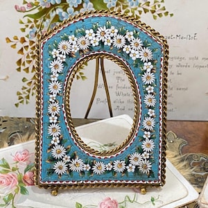 May include: An antique picture frame with a mosaic design. The frame has a teal background with white daisy-like flowers. The frame is trimmed with a gold-coloured metal and has a stand on the back.
