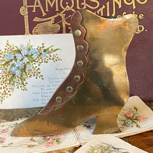 Könnte beinhalten: Ein Vintage-Stiefelobjekt aus Messing mit kupferfarbenem Besatz und dekorativen Nieten. Der Stiefel wird mit antiken Postkarten und einem Buch mit dem Titel "Famous Things of the World" präsentiert.