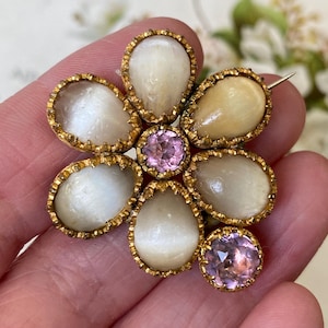 May include: A vintage flower brooch with a gold-tone setting. The brooch features six pear-shaped, off-white stones and two round, pink gemstones. The brooch is held in a hand, showcasing its intricate design and craftsmanship.
