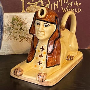 May include: A ceramic sphinx figurine in shades of tan, brown, and cream. The sphinx has a human head with a pharaoh's headdress and a lion's body. The figurine has a handle and is resting on a rectangular base.