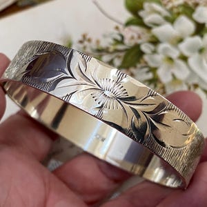 Puede incluir: Un brazalete plateado con un diseño floral grabado. El brazalete presenta una superficie texturizada y un tallado detallado de una flor y hojas. El fondo incluye flores blancas borrosas.
