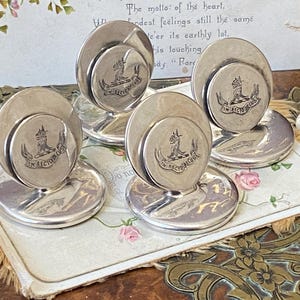 May include: A collection of four silver-coloured metal desk accessories. Each has a circular base and a raised disc with an emblem and the text "IN RECTO DECUS". They are displayed on a floral-patterned surface, with a vintage paper background.