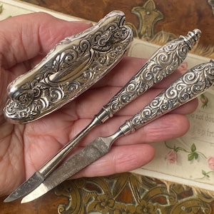 Op de afbeelding: Een antieke zilveren manicure set met een nagelknipper en twee gereedschappen. De set heeft sierlijke krullen op de handvatten en de koffer. De gereedschappen zijn gemaakt van zilver en staal. Waarschijnlijk uit het Victoriaanse tijdperk.