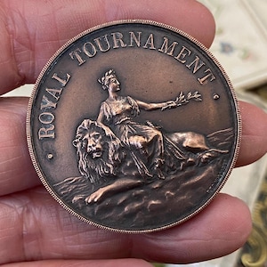 May include: A copper-coloured circular medal with the words "ROYAL TOURNAMENT" around the top edge. It depicts a seated woman holding a laurel branch, resting on a lion. The medal has a detailed, antique design.