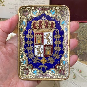 Può includere: Una scatola rettangolare smaltata antica con una cornice color oro. La scatola presenta uno sfondo blu con un dettagliato stemma, una corona e motivi floreali e geometrici decorativi in rosso, bianco e oro. La scatola è tenuta in una mano.