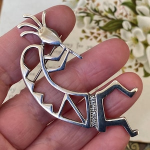Puede incluir: Un broche Kokopelli plateado, un símbolo nativo americano, se muestra. El broche presenta la figura de Kokopelli tocando una flauta, con un cuerpo curvo y piernas estilizadas. El broche se sostiene en una mano, con un fondo floral borroso.