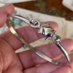 Könnte beinhalten: Ein silberfarbenes Armreif-Armband mit einem froschförmigen Verschluss. Das Armband hat ein einfaches, abgerundetes Band und ein detailliertes Froschdesign am Verschluss. Der Froschverschluss ist aufwendig geformt und verleiht dem Schmuck eine einzigartige Note.