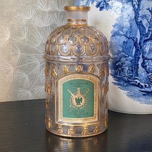 Puede incluir: Un frasco de perfume de vidrio transparente con detalles dorados y una etiqueta verde. El frasco tiene una parte superior abovedada con un borde dorado y está adornado con abejas doradas. La etiqueta dice "Eau de Cologne Imperiale" y "Guerlain".