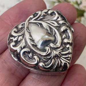 Puede incluir: Una caja de plata en forma de corazón con un intrincado patrón en relieve de hojas y volutas en la tapa. La superficie metálica tiene un acabado pulido con áreas hundidas oscurecidas que resaltan el detallado diseño de estilo floral.