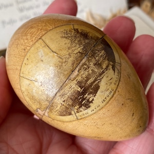May include: A vintage wooden egg with a sepia landscape image. The image is segmented and cracked, with the text "VIEW ON THE IRTHING GILS" visible. The egg is held in a hand. The object is a light, natural wood colour.