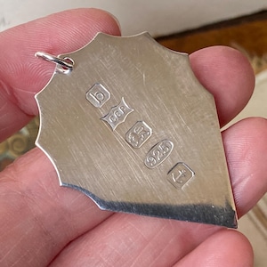 Può includere: Ciondolo in argento a forma di scudo con un piccolo anello nella parte superiore. Il ciondolo presenta diversi marchi, tra cui "925" che indica argento sterling. La superficie è lucida e riflettente.