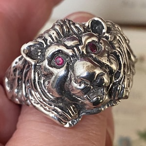 Può includere: Un anello color argento con la testa di un leone dettagliata. Il leone ha una criniera strutturata e due occhi di gemme rosse. Un gioiello audace.