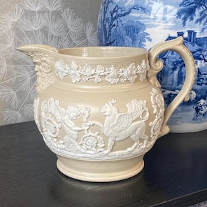 Puede incluir: Una jarra de cerámica beige con un diseño de relieve blanco detallado. La jarra presenta un asa, un pico y un relieve decorativo de figuras y criaturas míticas. Una pieza clásica de decoración del hogar.