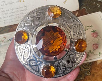 May include: A silver-toned brooch with a large, faceted amber-colored gemstone at the center, surrounded by four smaller gemstones. The brooch features Celtic knot and thistle designs.