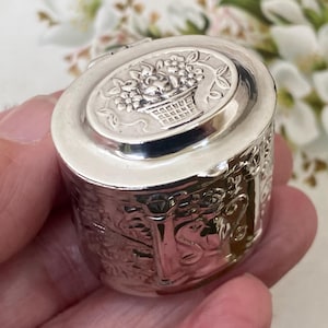 Puede incluir: Una pequeña caja de metal plateado de forma ovalada con tapa abatible. La parte superior de la tapa presenta un diseño en relieve de una cesta llena de flores. Los lados de la caja están decorados con intrincados patrones florales y volutas.
