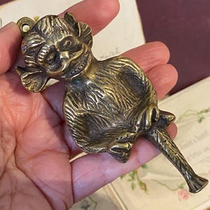 Puede incluir: Un llamador de puerta de latón vintage con forma de mono. El mono tiene detalles de pelaje y rasgos faciales, con un anillo para colgarlo. El llamador tiene un mango largo y cónico.