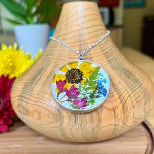 May include: A silver pendant necklace featuring a round, clear resin pendant with a pressed flower design. The pendant includes a yellow sunflower, pink and purple flowers, and blue forget-me-nots. The necklace is displayed on a wooden stand.