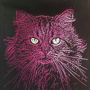 Puede incluir: Bolso tote negro con un diseño de cara de gato rosa. El pelaje del gato está representado con líneas texturizadas, y sus ojos son de un blanco brillante. El diseño está centrado en la bolsa, creando un fuerte contraste con el fondo negro. Los bigotes del gato son largos y prominentes.