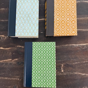 May include: Three hardcover books with decorative covers. One book has a light blue cover with a diamond pattern, another has an orange cover with a floral design, and the third has a green cover with a white floral pattern. The spines are dark blue or black.
