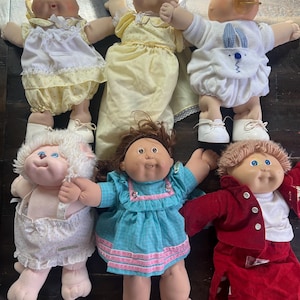 May include: Five Cabbage Patch Kids dolls in various outfits. The dolls are wearing a yellow dress, a white dress with a bunny design, a white dress with a pink and blue pattern, a white dress with a pink and blue pattern, and a red jacket with a white shirt and a red skirt.
