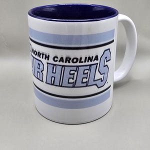 May include: White ceramic mug with a blue interior and a white handle. The mug features the words "North Carolina" above "HEELS" in light blue block letters. A blue stripe accents the top and bottom.