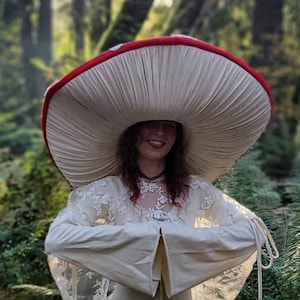 May include: A person wearing a large white mushroom hat with a red rim. The hat is very large and covers the person's head and shoulders. The person is wearing a white dress with lace details. The background is a forest.