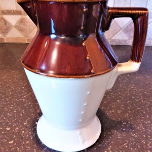 May include: A brown and white ceramic pitcher with a handle. The pitcher has a wide mouth and a rounded base.