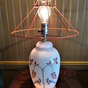 May include: A white ceramic table lamp with a painted design of two birds on a branch with red leaves. The lamp has a peach-colored wire cage shade.