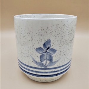 Op de afbeelding: Witte keramische bloempot met een gespikkelde afwerking en een blauw bloemenontwerp. De pot heeft drie horizontale blauwe strepen en een blauwe bloem met bladeren. Ideaal voor kamerplanten.