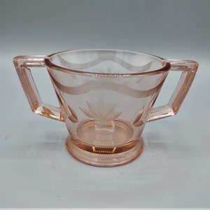 May include: A pink glass sugar bowl with two handles. The bowl has a delicate etched design.