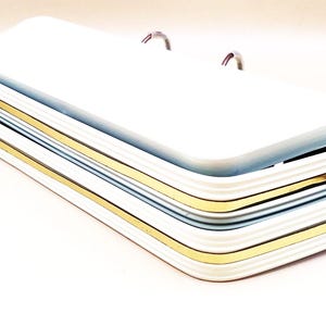 May include: Stack of three white rectangular serving trays with gold trim. The trays have a ridged design and are stacked on top of each other. Two small silver handles are visible on the top tray.
