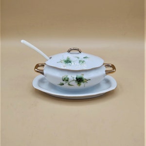 Pode incluir: Uma molheira de porcelana branca com tampa, acabamento dourado e pires a condizer. A molheira apresenta um desenho de folhas verdes e uma colher branca repousa na lateral. Este elegante prato de servir é perfeito para ocasiões especiais.