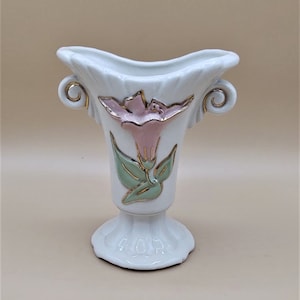 以下が含まれることがあります： ピンクとグリーンの花柄の白いセラミックの花瓶。花瓶は金色のアクセントと2つのハンドルが付いています。