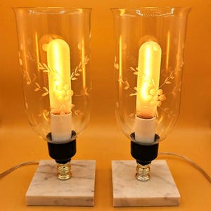 Puede incluir: Dos lámparas de mesa vintage con pantallas de vidrio transparente grabadas con diseños florales. Cada lámpara presenta una bombilla larga y amarilla, una base negra y un acento dorado. Las lámparas se asientan sobre bases cuadradas de mármol blanco.