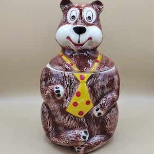 May include: A brown ceramic cookie jar shaped like a bear wearing a yellow tie with red polka dots. The bear has a smiling face with black eyes and a black nose.